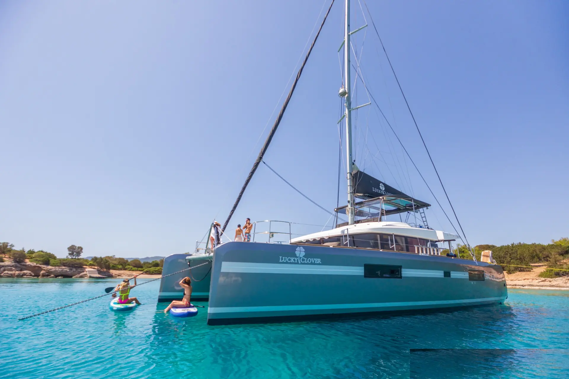 Lucky Clover at anchor in a turquoise bay, the tender alongside and two paddle boards in the water